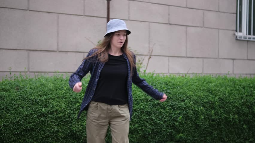 young woman in a blue plaid shirt and beige pants dances hip-hop in front of a tall green hedge and a beige brick wall. She wears a white bucket hat and black shirt, with her long hair flowing freely