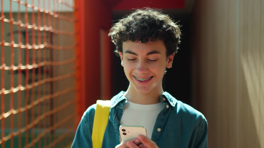 Happy smiling positive Caucasian teen boy student guy teenager man male son pupil in university school corridor holding mobile phone addict social media good news smile toothy braces education study