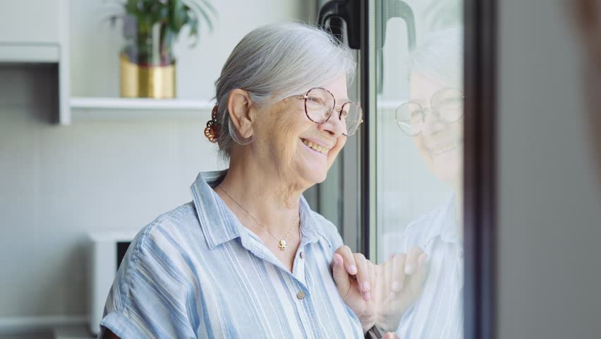 Dreamy smiling elderly 60s hoary woman looking in distance out of window, recollecting good memories or visualizing future at home, planning vacation or enjoying peaceful mindful day alone indoors.
