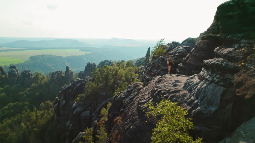 Woman hiking on the edge of a rocky cliff in Schrammsteine Elbe Sandstone Mountains in Germany with forest and hills in the background
