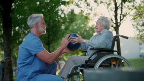 Male caregiver exercising with elderly patient on wheelchair, using small exercise ball. Geriatric physiotherapist during outdoor workout therapy in public park, nursing home garden. - Powered by Shutterstock - Get 15% off with code: PIKWIZARD15