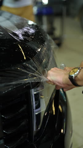 Closeup of auto service man applying transparent protective film to back door of black car, using squeegee to smooth out film and remove air bubbles. Process professional auto detailing, slow motion.