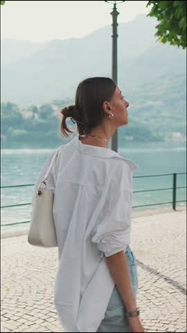 Vertical video, Young brown-haired woman dressed in white shirt, walking along embankment with lake and mountains in background