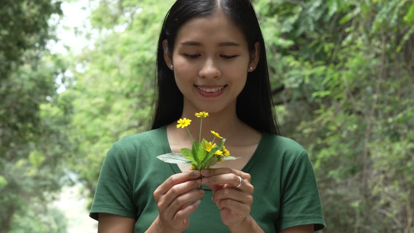 A cheerful young asian woman enjoys a sunny day outdoors, holding a vibrant bunch of yellow flowers. Smiling in a green field, she embodies the joy and freedom of summer in the countryside.