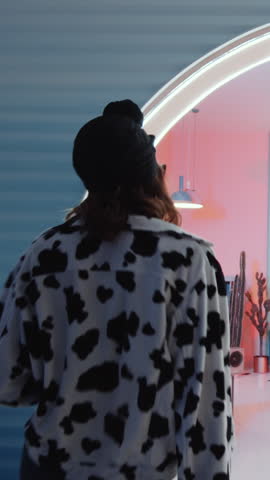 Vertical shot of trendy girl wearing sunglasses and cow print jacket walking into colorfully lit room with stylish interior, turning music on and performing expressive vogue dance