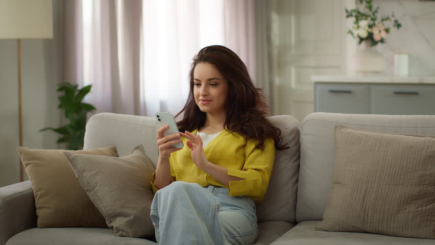 Happy family spending weekend together. Caucasian woman female mother using mobile cell phone online app active little girl child kid schoolgirl daughter jumping on sofa hugging mom inside living room