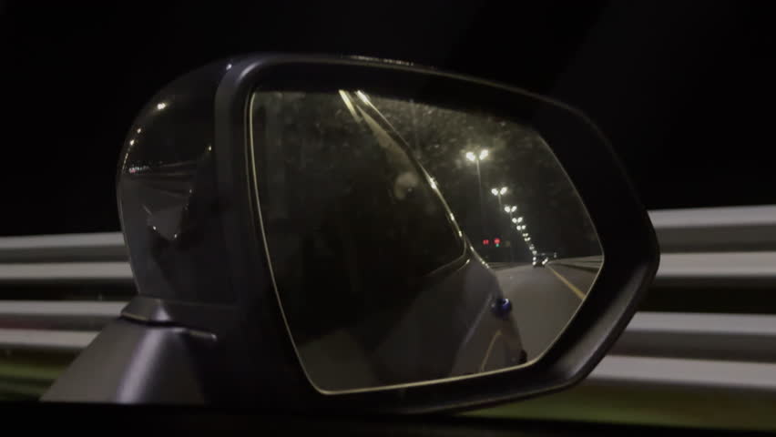 The reflection of night sky and road in rearview mirror of a car driving down a dark highway. Image is captured from right rear view mirror. Modern mirror of black car reflects passing street lights.