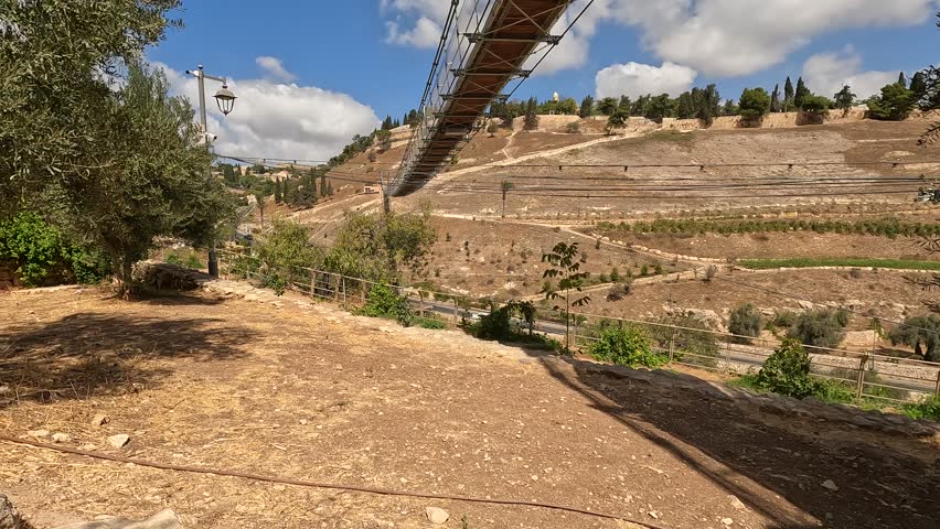 the suspension bridge over Gei Ben Hinnom near the Old City of Jerusalem, with the Mount of Olives and cloudy skies in the background. late summer