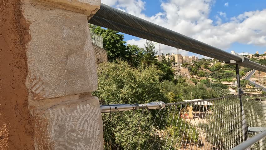walking on the suspension bridge over Gei Ben Hinnom near the Old City of Jerusalem, with the Mount of Olives and cloudy skies in the background.  late summer