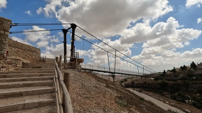 the suspension bridge over Gei Ben Hinnom near the Old City of Jerusalem, with the Mount of Olives and cloudy skies in the background. Perfect for projects on Israeli landscapes and historical sites.