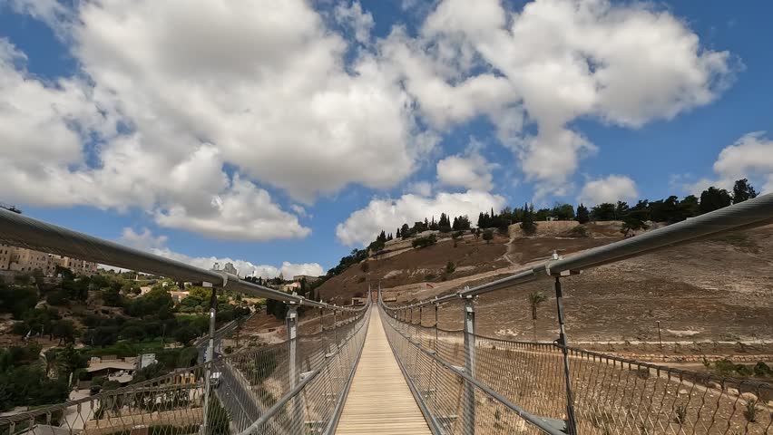 Walking on the suspension bridge over Gei Ben Hinnom near the Old City of Jerusalem, with the Mount of Olives and cloudy skies in the background. Israel