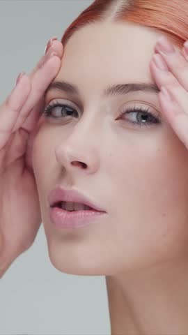 Vertical studio portrait of young, beautiful and natural redhead woman applying skin care cream. Face lifting, cosmetics and make-up concept.