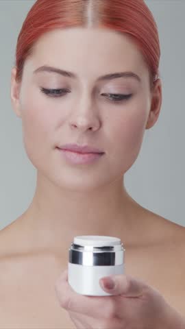Vertical studio portrait of young, beautiful and natural redhead woman applying skin care cream. Face lifting, cosmetics and make-up concept.