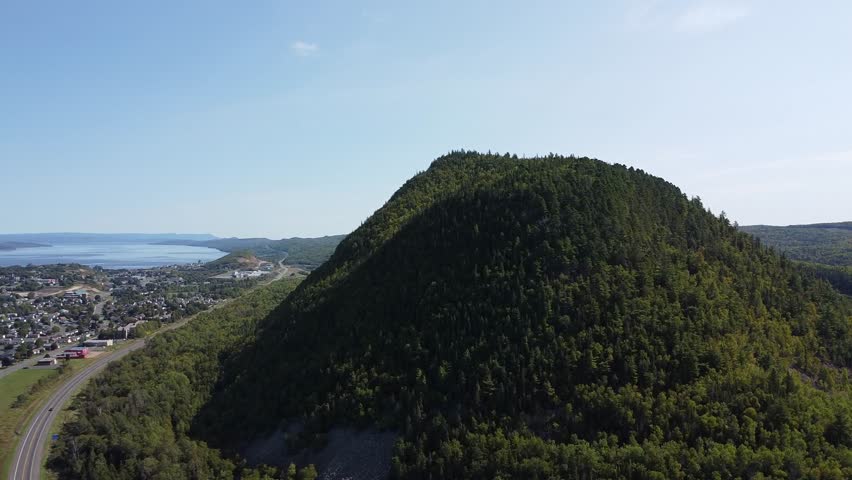 Aerial drone view of Sugarloaf Mountain, an ancient volcano in Atholville, New Brunswick. The footage shows the forested peak with the city of Campbellton visible in the background under a clear blue sky. Captured in Canada in 2024.