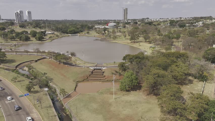 Drone flies over highway into park with large brown lake in Campo Grande, Mato Grosso do Sul, Brazil