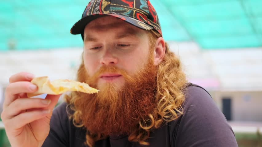 Smiling hungry red-bearded man bites off a piece of pizza squints with pleasure fast food lover snacking outdoors in city cafe.