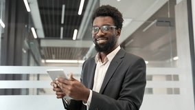 Smiling businessman holding digital tablet in modern business office. Confident black entrepreneur in a formal suit with glasses, looking at camera. Headshot of a successful professional manager - Powered by Shutterstock - Get 15% off with code: PIKWIZARD15