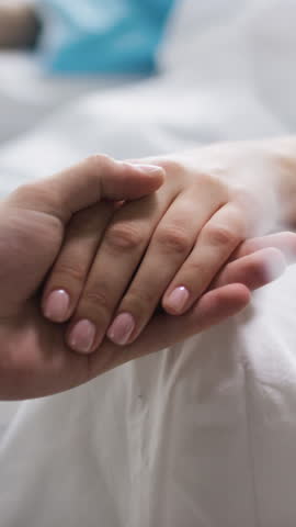 Vertical close-up of unrecognizable man holding hand of anonymous sick woman lying in hospital bed or in palliative care and gently stroking her manicured fingers