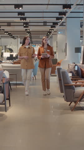 Full vertical shot of saleswoman with badge and young Caucasian female customer walking around furniture showroom, looking at couches and sofas, discussing and planning purchase for new apartment