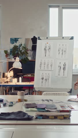 Full vertical shot of young creative African American clothing designer presenting garment sketches for new fashion collection to team at meeting at clothing factory or atelier