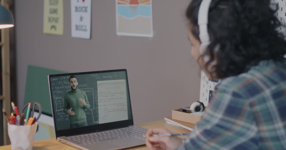 Teenage boy watching online lesson on laptop screen writing in notebook studying in internet from home. E-learning and modern education concept.