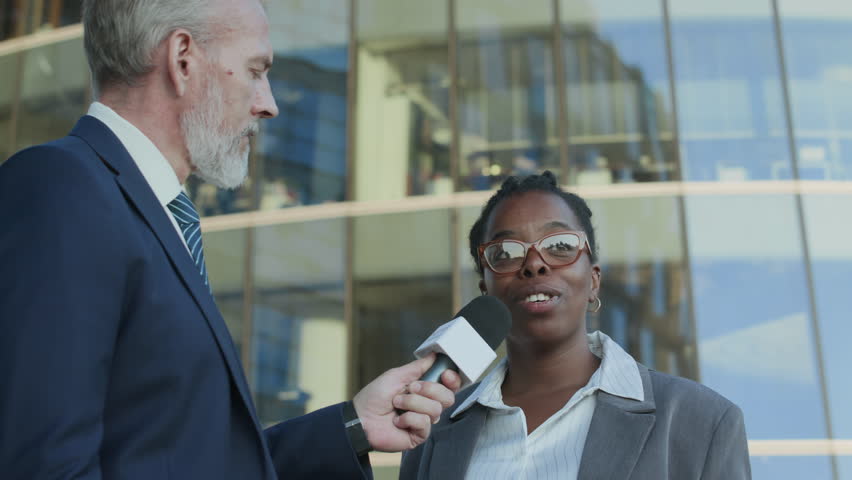 Low angle arc shot of young Black female entrepreneur giving interview to news channel reporter while standing in front of tall glass office building in downtown on sunny day