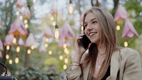 An attractive young woman chats on her mobile phone while seated at a decorated, illuminated outdoor cafe. The setting exudes a cozy, inviting atmosphere. Young Woman Making a Phone Call Outdoor Cafe - Powered by Shutterstock - Get 15% off with code: PIKWIZARD15