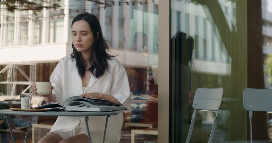 A young woman is seen sipping coffee while engrossed in a book at an outdoor cafe. This image captures a moment of relaxation and tranquility in an urban setting, highlighting a leisurely lifestyle.