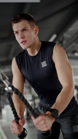 A determined young man trains intensely in a gym using battle ropes, showcasing effort and fitness focus. Focused Young Man Working Out with Battle Ropes in a Gym. Vertical video.
