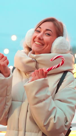 Smiling middle-aged woman in warm winter jacket and white fur earmuffs holds Christmas candies in her hands stands at a festive holiday market, surrounded by Christmas lights and joyful atmosphere