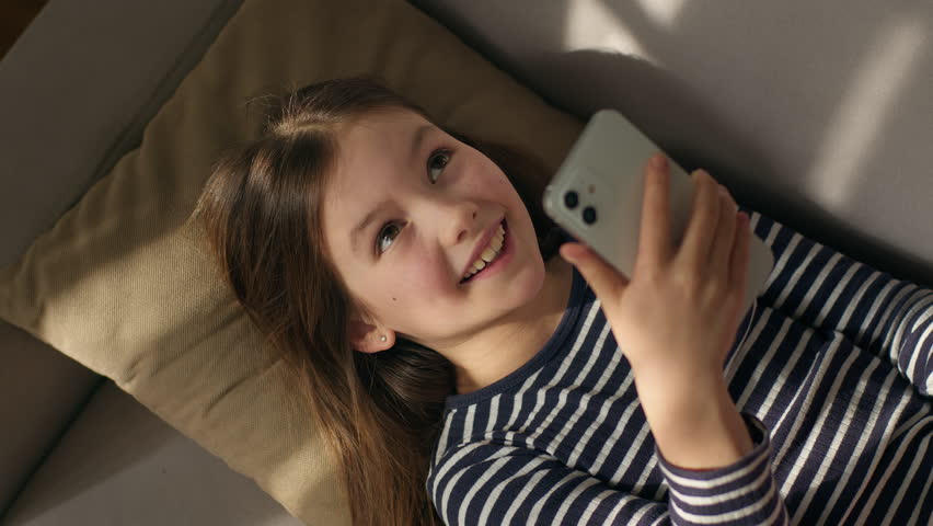 Caucasian European little girl lying on sofa holding using mobile phone smartphone child gadget online texting sms social networks smiling laughing enjoyment watching funny video internet chatting kid