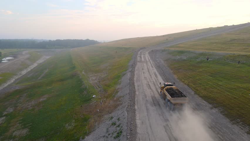 Heavy dump truck driving up dirt road to landfill during sunset, aerial view