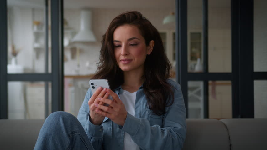 Caucasian woman user girl female housewife homeowner at home apartment couch sofa using mobile phone smartphone app browsing web service social media internet online shopping watching video cellphone