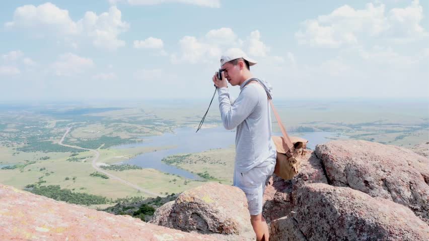 Male photographer wearing camera bag taking photos with a film camera at Mount Scott, Oklahoma facing idyllic valley. -wide static shot
