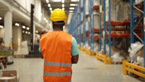 Rear view of an african american male warehouse worker wearing a safety vest and hardhat is walking through a busy warehouse, looking around at the inventory. The video is in slow motion - Powered by Shutterstock - Get 15% off with code: PIKWIZARD15