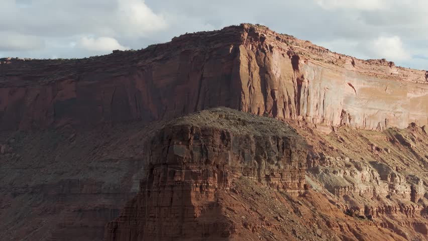 A 70mm high-flying orbital drone shot around a massive, isolated rock tower, with an even bigger intensely contoured cliff face behind it, in the unique and extreme desert landscape of Moab, Utah.