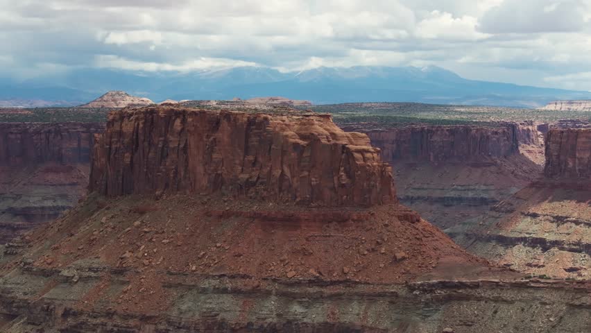 A high-flying drone shot of the unique and extreme desert landscape of Moab, Utah. The camera orbits around a massive natural rock structure over the canyon, with white-top mountains in the distance.