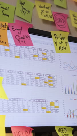 Vertical video. In a workspace filled with colorful papers, a young man attaches bright sticky notes to his computer screen, emphasizing time organization and effective planning
