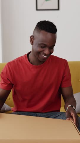 Vertical HD video of happy african man opening cardboard box at home. Young millennial male sitting on sofa unpacking parcel delivery box of new clothes. Online shopping shipment concept