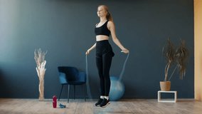 A woman in athletic wear jumps rope inside a bright fitness room. The space features a blue wall, plants, and exercise equipment, creating an energetic atmosphere for her workout. - Powered by Shutterstock - Get 15% off with code: PIKWIZARD15