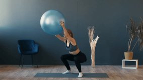 A woman is executing squats while holding a stability ball in a beautifully decorated indoor space. The setting features a chair and decorative plants, creating a serene atmosphere. - Powered by Shutterstock - Get 15% off with code: PIKWIZARD15