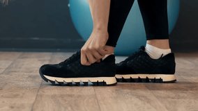 A person kneels and ties their shoelaces on a pair of black athletic sneakers while preparing for a workout in a modern gym. The environment features a wooden floor and blue exercise ball nearby. - Powered by Shutterstock - Get 15% off with code: PIKWIZARD15