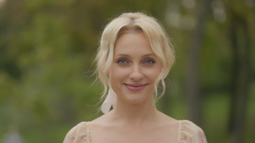 A close-up portrait of a blonde middle-aged woman, smiling warmly and relaxed outdoors in natural sunlight.