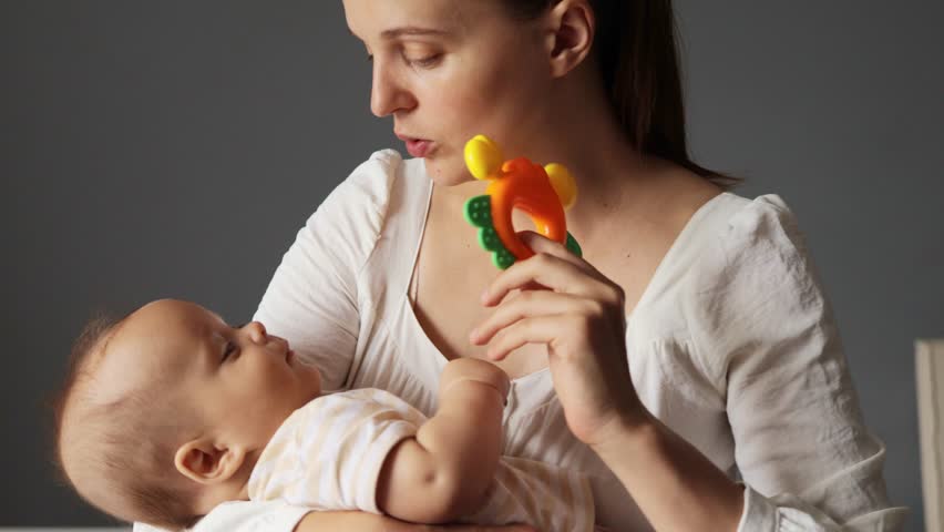 Good looking Caucasian woman wearing white dress playing with her infant baby showing toy to her daughter or son rocking calming down child at home
