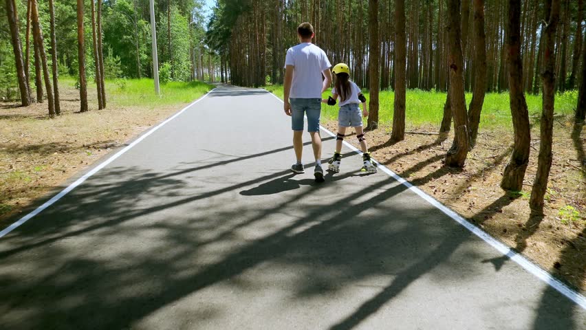 Father skating with her daughter in park. Young smiling parent enjoying free time with her little girl, roller skating outside. Happy cute child wearing a helmet and bonding with man