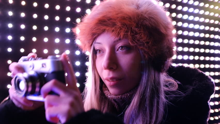 Woman with a hat taking photos with a retro camera on a winter night by the neon lights