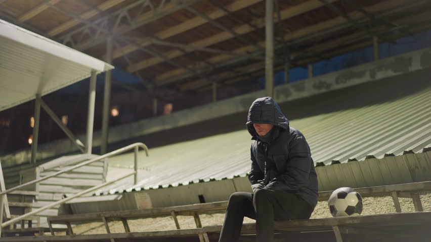 A man dressed in a black outfit and jacket sits alone in a quiet stadium with his hands clasped between his legs and a ball close to him slightly moved by the wind