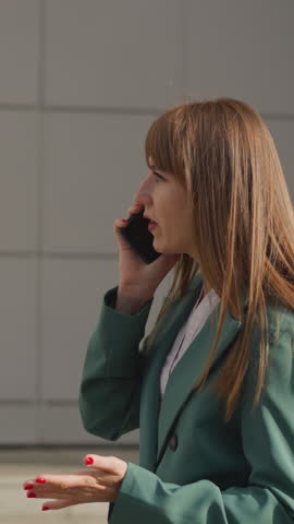 Displeased woman communicates on smartphone waving hand emotionally. Redheaded lady discusses problematic issues walking down street in big city closeup side view