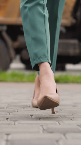 Stylish woman in high heel shoes walks on paving slabs road against dump truck standing on road. Elegant lady in green suit goes to work closeup slow motion