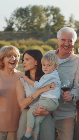 Vertical group portrait of cheerful grandparents, their daughter and little grandson posing for camera with wine glasses while having family party outdoors